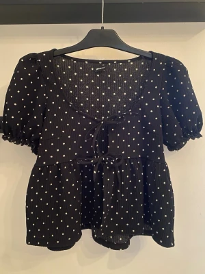 Svart prickig blus med knyt - Supersöt svart blus från Gina Tricot med vita prickar, puffärmar och två knyt framtill. Blusen har en djup rundringning och lätt volang nedtill. 