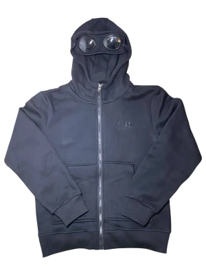 Svart hoodie med glasögon från C.P. Company - Svart hoodie från C.P. Company med hel dragkedja, ribbade muddar och två fickor framtill. Huvan har inbyggda glasögon, vilket ger en unik och futuristisk look. Tillverkad i mjukt bomullsmaterial och har diskret logga broderad på bröstet.