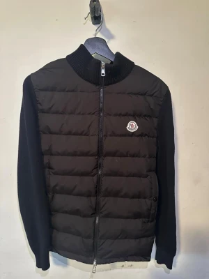Svart cardiganjacka från Moncler - Snygg svart cardiganjacka från Moncler med quiltad framsida och stickade ärmar. Jackan har ribbad krage, dragkedja framtill och Moncler-logga på bröstet. Använde aldrig de då den inte passade mig så den är helt ny. Storlek S/M
