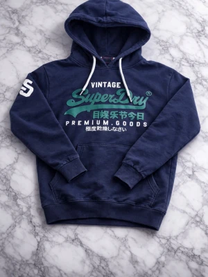 Mörkblå Superdry hoodie med tryck - Snygg mörkblå hoodie från Superdry med stort grönt och vitt tryck på bröstet och japanska tecken. Klassisk känguruficka, vita dragsnören i huvan och tryckt siffra på ena ärmen. Perfekt för en avslappnad streetwear-look.