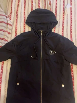 Svart vindjacka från Trapstar - Snygg svart vindjacka från Trapstar med huva och dragkedja framtill. Jackan har två fickor med tryckknappar och ett Trapstar-märke på bröstet. På ryggen finns en stor Trapstar-logga i vit text. Perfekt för streetwear-stilen.