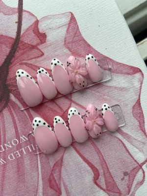 Rosa press-on naglar med blommor - Snygga press-on naglar i ljusrosa med vita franska tippar och svarta prickar. Några naglar har 3D-blommor i rosa med guld detalj i mitten. Ovala och mandelformade, perfekta för dig som vill ha en söt och trendig look.