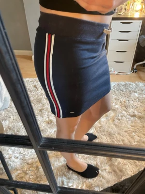 Marinblå kjol med ränder Tommy Hilfiger - Sportig marinblå kortkjol från Tommy Hilfiger med vita och röda ränder längs sidorna. Kjolen har bred resår i midjan med snörning och är tillverkad i mjuk bomull. Perfekt för en avslappnad och trendig look. Sitter som en tajt S