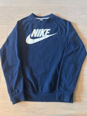 Mörkblå sweatshirt från Nike - Snygg mörkblå sweatshirt från Nike med stort vitt logotryck på bröstet. Tröjan har rund halsringning, långa ärmar och ribbade muddar vid ärmslut och nederkant. Perfekt för en sportig och avslappnad stil.