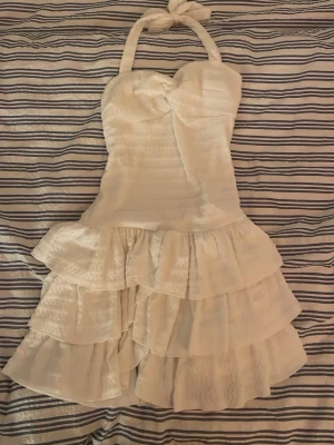 Vit volangbaddräkt med halterneck - Supersöt vit baddräkt med halterneck och knytband i nacken. Baddräkten har flera lager volanger nedtill och en snygg twistdetalj vid bysten. Materialet ser ut att vara stretchigt och bekvämt, perfekt för strandhäng eller poolparty.