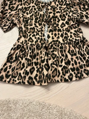 Leopardmönstrad blus med knyt - Säljer en leopardmönstrad topp med rosetter samt volanger nedtill. En perfekt topp till dig eller din dotter som vill ha en skön men fin topp. Toppen är nästan helt ny och säljer pga för liten storlek. Kontakta gärna vid intresse. Storlek 146/152