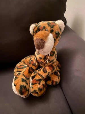 Mjuk leopard gosedjur - Säljer ett mjukt gosedjur i form av en leopard med brun, svart och orange fläckig päls. Nosen är brun och vit, tassarna är vita och den har långa ben och svans. Perfekt som mysig inredningsdetalj eller present till någon som gillar djur.
