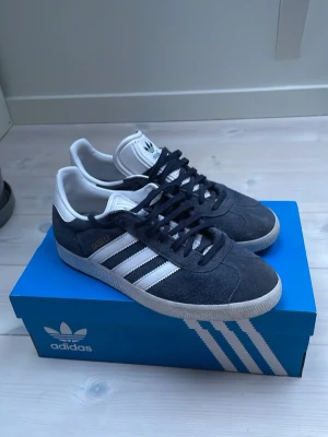 Adidas Gazelle mörkblå sneakers - Hej, jag säljer nu dessa snygga Adidas Gazelle sneakers i mörkblå mocka med vita detaljer och klassiska tre ränder på sidan. Skorna har vit sula och vit hälkappa samt guldfärgad Gazelle-text på sidan. Perfekta för dig som letar efter ett par stilrena sneakers till sommaren. Skorna är size 40 2/3 men passar dig som har size 41. De är i bra skick bortsätt från att de är slitna i hälen. Därav är priset lägre. Vid fundering tveka inte på att skriv.🕺