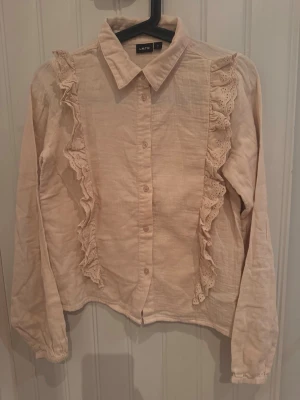 Beige blus med volang från NameIT - Supersöt beige blus från LMTD med långa ärmar och volangdetaljer framtill. Blusen har klassisk krage, knäppning hela vägen och fina spetsvolanger som ger en romantisk touch. Perfekt att styla med jeans eller kjol för en trendig look.