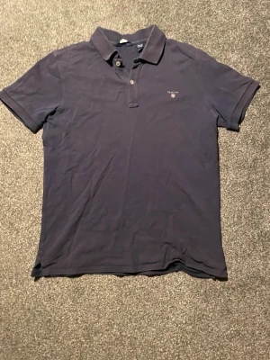 Mörkblå pikétröja från Gant - Klassisk mörkblå pikétröja från Gant med korta ärmar och krage. Tröjan har två knappar vid halsen och en liten broderad logga på bröstet. Perfekt för en stilren och avslappnad look.