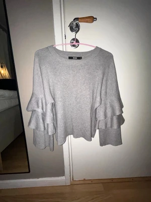 Grå stickad tröja med volangärmar - ‼️PRIS KAN DISKUTERAS‼️Säljer en ljusgrå stickad tröja från Bik Bok med trendiga volangärmar i två lager. Tröjan har rund halsringning och en avslappnad passform som ger en cool och modern look. Perfekt att styla till både jeans och kjol.