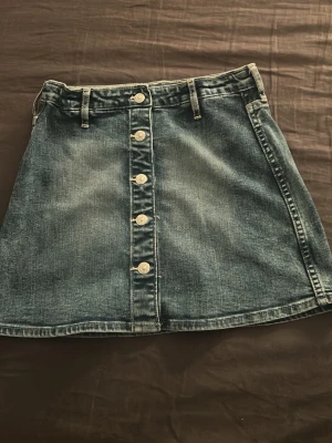 Blå jeanskjol med knappar framtill - Säljer en klassisk blå jeanskjol i kort modell med knappar längs hela framsidan. Kjolen har bälteshällor och en lätt utställd passform. Perfekt till vardag och när man vill ha lite fest kläder!❣️