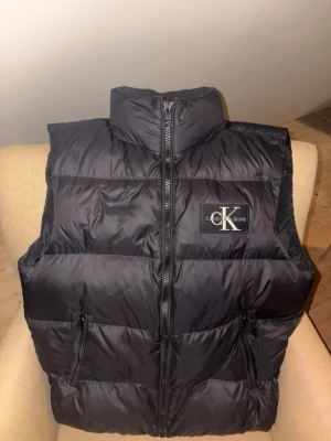 Svart dunväst från Calvin Klein  - Snygg svart dunväst från Calvin Klein  med hög krage och dragkedja framtill. Västen har två praktiska fickor med dragkedja och en tydlig CK-logga på bröstet. Perfekt för lager på lager och streetwear-stil.