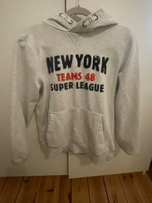 Grå hoodie med New York-tryck - Ljusgrå hoodie med huva och snörning. Tryck på bröstet med texten 'NEW YORK TEAMS 48 SUPER LEAGUE' i svart och rött. Stor känguruficka framtill och mjukt material, perfekt för chill dagar.