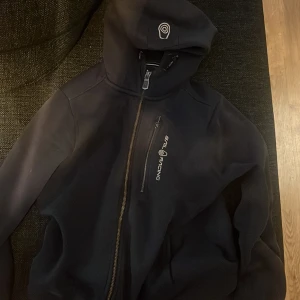 Svart vindjacka från Sail Racing - Mörkblå hoddie  från Sail Racing med huva och snyggt logotryck på bröstet och huvan. Hoddien  har en bröstficka med dragkedja och grov dragkedja framtill. Materialet är mjukt och känns sportigt, perfekt för dig som gillar stilren design och funktion.