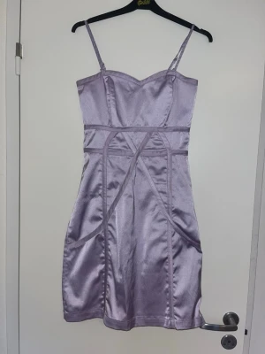 Ljuslila satin klänning med smala band - Säljer en snygg ljuslila kort klänning i satin med smala axelband och figurnära passform. Klänningen har markerade sömmar och en lätt glansig yta som ger en elegant look. Perfekt för dig som vill sticka ut med en och stilren outfit.