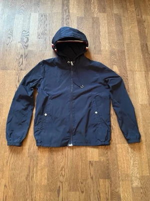  Moncler vinjacka - Snygg mörkblå vindjacka från Moncler med huva och dragsko. Jackan har två framfickor med tryckknappar och dragkedja framtill. Tillverkad i lätt och tåligt material, perfekt för blåsiga dagar. Moncler-logga på ärmen ger en exklusiv touch.