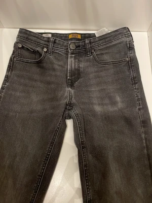 Grå slim fit jeans Jack & Jones - Snygga grå slim fit jeans från Jack & Jones, modell Glenn. Jeansen har klassisk femficksdesign, knappgylf och subtila slitningar för en trendig look. Perfekta för dig som gillar en smalare passform och vill ha ett par stilrena jeans i garderoben.
