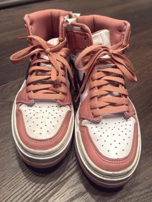 Nike Air Jordan 1 High rosa/vit - Säljer ett par Nike Air Jordan 1 High sneakers i rosa och vitt med svart swoosh. Skorna har högt skaft, perforerad tåbox och klassisk snörning. Tillverkade i läder med ikoniska detaljer på sidan och tungan. Perfekta för dig som gillar streetwear och snygg design.