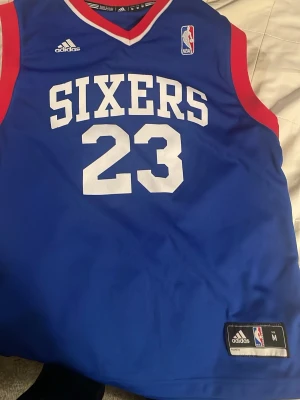Sixers basketlinne Williams #23 Adidas - Blått basketlinne från Adidas med röda detaljer och vita tryck. Framsidan har texten 'SIXERS' och nummer 23, baksidan har namnet 'WILLIAMS'. Officiell NBA-logga på bröstet och etikett nertill. Perfekt för basketfans och träning.