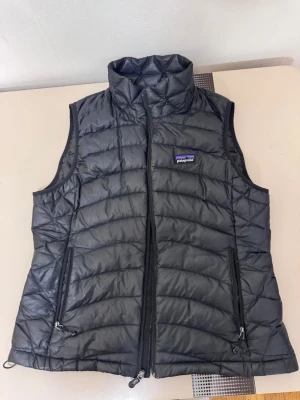 Svart dunväst från Patagonia - Snygg svart dunväst från Patagonia med hög krage och två dragkedjeförsedda fickor framtill. Västen har quiltad design och diskret logga på bröstet. Perfekt för lager-på-lager och håller dig varm under kyliga dagar.  Den har ett jätte litet hål i nacken sen måste man stänga dragkedjan på ett speciellt för att den ska stängas. 