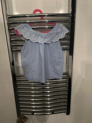 Blåvit randig ärmlös blus med spets - Söt blåvit randig ärmlös blus med bred, vit spetskrage och rund hals. Blusen har tunna vertikala ränder och en luftig passform, perfekt för varma dagar. Spetsdetaljen ger en romantisk touch till toppen.