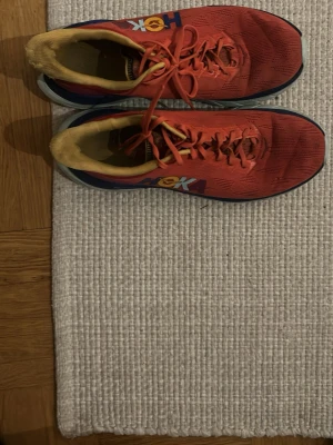 Röda Hoka sneakers med mesh - Säljer ett par röda Hoka sneakers med ventilerande mesh-ovandel, gula detaljer på insidan och logga i blått och gult på sidan. Skorna har klassisk snörning och en låg siluett med bekväm sula som passar perfekt för dig som gillar sportig stil.