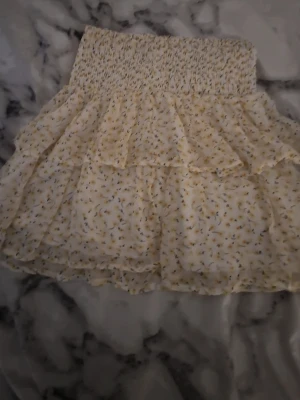 Blommig volangkjol i ljusgult - Supersöt kort kjol med volanger i ljusgul ton och små blommor. Kjolen har bred smock i midjan för skön passform och är gjord i ett lätt, luftigt material som passar perfekt till varma dagar.