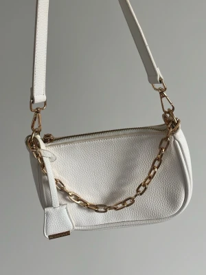 Vit axelremsväska med guldkedja - Stilren vit handväska i strukturpräglat material med justerbar axelrem och dekorativ guldfärgad kedja. Väskan har dragkedja upptill och en liten tag med metallogo. Perfekt accessoar för en trendig look.