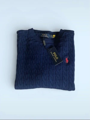 Mörkblå kabelstickad tröja Polo Ralph Lauren - Snygg mörkblå kabelstickad tröja från Polo Ralph Lauren med rund halsringning och röd broderad logga på bröstet. Tröjan har långa ärmar och ribbade muddar. Perfekt för dig som gillar klassisk och stilren design.