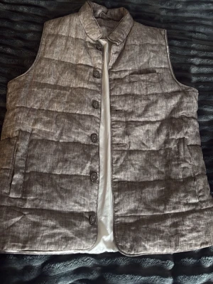 Beige dunväst från John Henric - Snygg beige/brun dunväst från John Henric med ståkrage och knäppning med fem knappar framtill. Västen har två sidofickor och en bröstficka, quiltad design och ett stilrent uttryck. Perfekt för lager på lager och enkel att matcha med olika outfits.