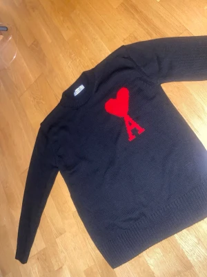 Svart stickad tröja från Ami Paris - Svart stickad tröja från Ami Paris med rött hjärta och bokstaven A på bröstet. Tröjan har rund halsringning och ribbade muddar vid ärmslut och nederkant. Perfekt för dig som gillar stilrena plagg med en twist.