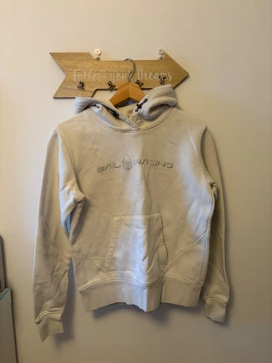 Vit hoodie från Sail Racing - Snygg vit hoodie från Sail Racing med broderad logga på bröstet. Hoddien har också en framficka framtill oxh en snygg luva. Perfekt för en sportig stil. Barnstorlek M. 