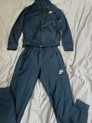 Blå Nike träningsset med dragkedja - Snyggt blått träningsset från Nike med matchande jacka och byxor. Jackan har hel dragkedja, huva och två fickor med dragkedja. Byxorna har resår i midjan och dragkedjeförsedda fickor. Båda delarna har Nike-logga i vitt. Perfekt för träning eller chill.