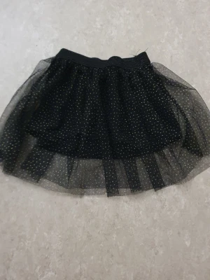 Kjol - Kort svart tyllkjol med små guldprickar och elastisk midja. Kjolen har flera lager för extra volym och en glittrig look som sticker ut. Perfekt för dig som vill ha något extra till outfiten.