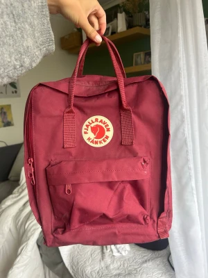 Vinröd Fjällräven Kånken ryggsäck - Klassisk vinröd Fjällräven Kånken ryggsäck med ikonisk rund logga framtill. Ryggsäcken har ett stort huvudfack, en dragkedjeförsedd ytterficka och två sidofickor. Tillverkad i slitstarkt syntetmaterial med bärhandtag och justerbara axelband. Väskan är aldrig använd men har ett litet slitet band