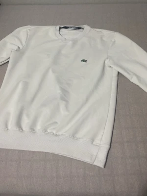 Vit sweatshirt/kofta från Lacoste - Klassisk vit sweatshirt från Lacoste med rund halsringning och den ikoniska gröna krokodilloggan broderad på bröstet. Tröjan har långa ärmar och ribbade muddar vid ärmslut och nederkant. Perfekt för en clean och stilren look. Pris kan diskuteras så hör av dig. 