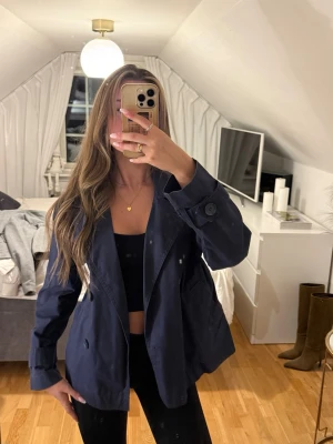 Mörkblå trenchcoat med stora slag - Snygg mörkblå trenchcoat med dubbelknäppning och stora slag. Jackan har breda ärmar med dekorativa knappar vid ärmsluten och en avslappnad passform. Perfekt för dig som vill ha en klassisk men modern look. Materialet känns mjukt och följsamt.