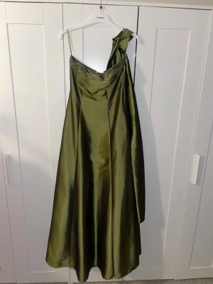 Grön långklänning med pärlor - Elegant grön långklänning i glansigt tyg med en axelbandslös design och dekorativa pärlor längs överdelen. Snörning i ryggen för förbättrad passform och tillhörande sjal för att bära runt axlarna. Perfekt för speciella tillfällen.