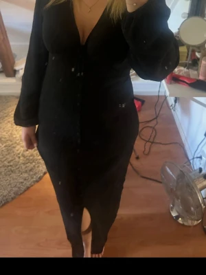 Svart långklänning med knappar - Svart långklänning med v-ringning och långa ärmar. Klänningen har knappar framtill och en liten slits nedtill. Materialet ser ut att vara polyester med en lätt struktur. Enkel och stilren design som passar till många tillfällen. En adoore liknande klänning 