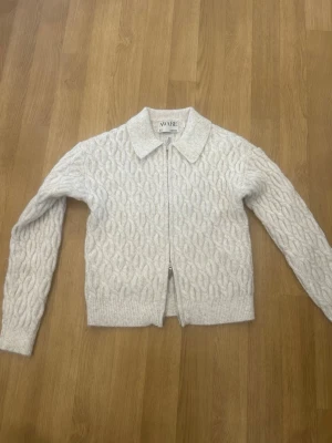 Vit kabelstickad kofta från AWARE - Kabelstickad vit kofta från AWARE med dragkedja framtill och klassisk krage. Koftan har långa ärmar och ribbade muddar vid ärmslut och nederkant. Perfekt för lager-på-lager och en stilren look.