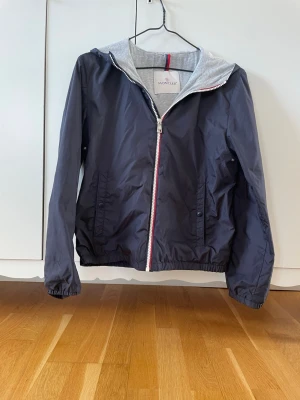 Mörkblå vindjacka från Moncler - Snygg mörkblå vindjacka från Moncler med grå huva och dragkedja med röd, vit och blå detalj. Jackan har två fickor med knappstängning, elastiska muddar och en sportig look. Perfekt för blåsiga dagar och enkel att matcha till olika outfits. kontakta för funderingar