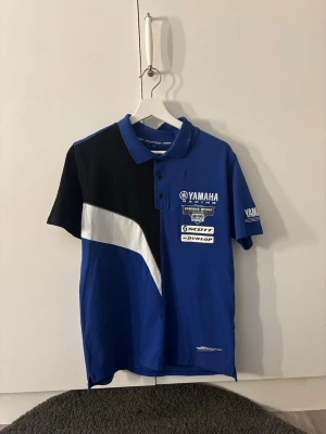 Blå Yamaha Racing piké t-shirt - Snygg blå Yamaha Racing piké t-shirt med svart och vit detalj på framsidan samt tryckta loggor från Yamaha, Scott och Dunlop. T-shirten har krage med knappar och korta ärmar. Perfekt för dig som gillar motorsport och vill ha en sportig look. Storlek: M och i ett nytt skick. Lägg ett bud, vill bli av med! 