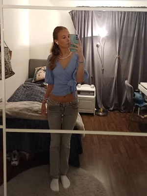 Blå omlottopp med knyt - Säljer en ljusblå omlottopp med knyt i midjan och v-ringning. Toppen har korta puffärmar och är croppad, vilket ger en trendig och avslappnad look. Storlek M men passar även S