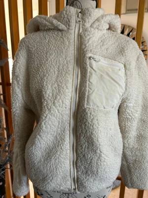 Vit teddyjacka med huva och ficka - Mysig vit teddyjacka med dragkedja framtill och huva. Jackan har en praktisk bröstficka i nylon med dragkedja som detalj. Tillverkad i fluffigt teddy-material som ger en avslappnad och trendig look. Perfekt för kyliga dagar när du vill vara både varm och snygg.