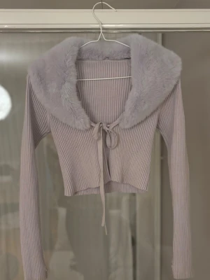 Ljuslila ribbad kofta med fuskpäls - Trendig ljuslila ribbad kofta med stor fluffig fuskpäls-krage och knytband framtill. Croppad modell med långa ärmar, perfekt för att lyfta din outfit med en mjuk och lyxig känsla. Pälsen går att ta av och på med hjälp av knapparna som sitter på insidan.  