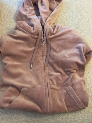 Rosa velour hoodie från Juicy Couture - Mjuk rosa hoodie i velour från Juicy Couture med dragkedja, fickor framtill och broderad logga på bröstet. Huva med dragsko och ribbade muddar vid ärmslut. Perfekt för en chill och trendig look.