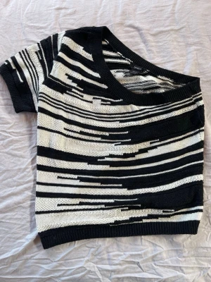 Svartvit oneshoulder topp med ränder - Snygg stickad topp i svart och vitt med abstrakt randigt mönster. Modellen är oneshoulder med kort ärm på ena sidan och bred ringning. Perfekt för dig som vill ha en unik och trendig look.Aldrig använd!