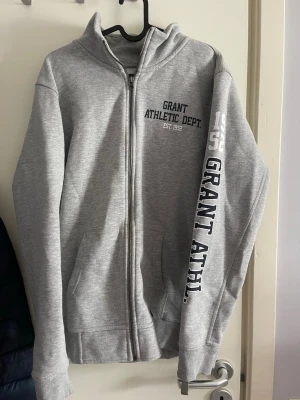 Grå zip hoodie Grant Athletic Dept. - Snygg grå zip hoodie från Grant Athletic Dept. med tryck på bröstet och ärmen. Hoodien har dragkedja, två fickor fram och en klassisk huva. Perfekt för en sportig och avslappnad stil. Tillverkad i mjukt sweatshirtmaterial.