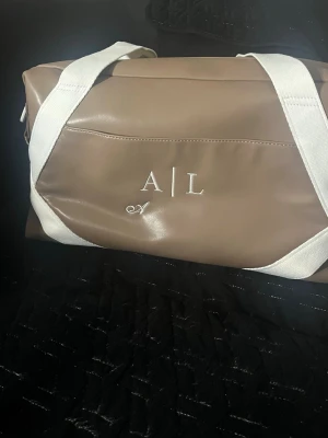 Weekendbag  - Brun väska från Pristine Collection ROFA DESIGN med vita detaljer och justerbar axelrem. Väskan har ett fack med dragkedja på insidan och en på baksidan. Väskan har guldfärgade detaljer. Den är oanvänd med lapparna kvar.   DUFFLE BAG (info från hemsidan)  I väskan hittar du: - Ett rymligt huvudfack - Ett innerfack med dragkedja - Två fickor för förvaring av t.ex. mobil och nycklar - Två ytterfack för mindre förvaring  Märkmetod: Brodyr Längd: 43 cm Höjd: 23 cm Djup: 18 cm Material: Veganläder/Bo